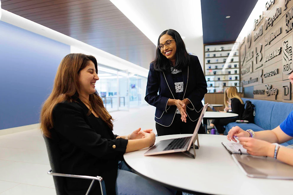 A Creighton University faculty member smiling and talking with a student using a laptop in a modern campus setting.