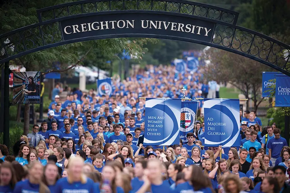 Students in blue shirts walking under the Creighton University archway during a campus event, holding banners that read "Men and Women for and with Others" and "Ad Majorem Dei Gloriam."