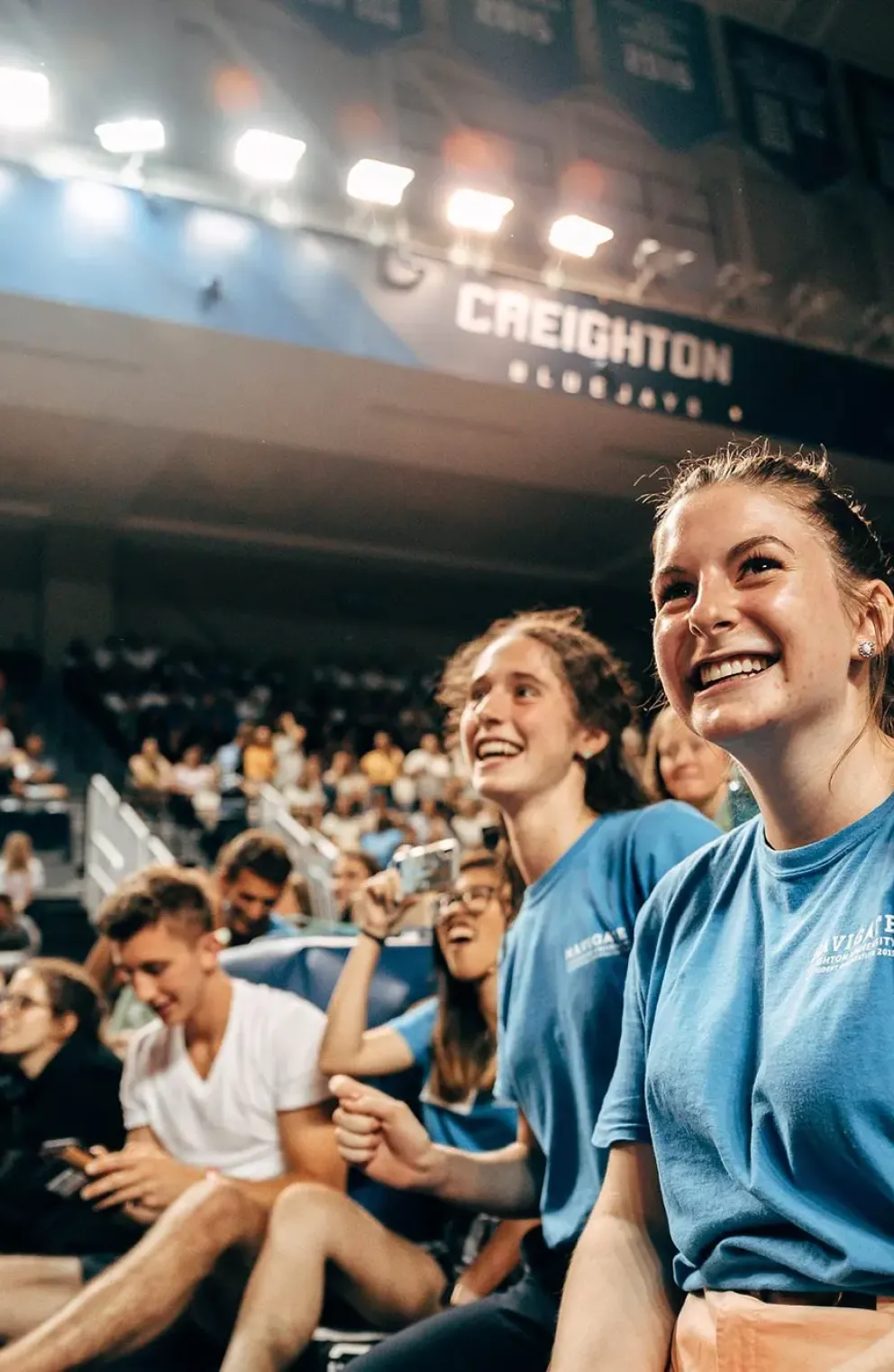 Smiling students in blue shirts watching a Creighton University event surrounded by a crowd.