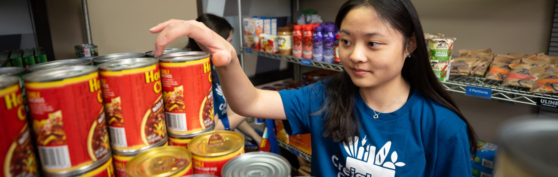 A student wearing a creighton cupboard tshirt putting canned goods on a shelf