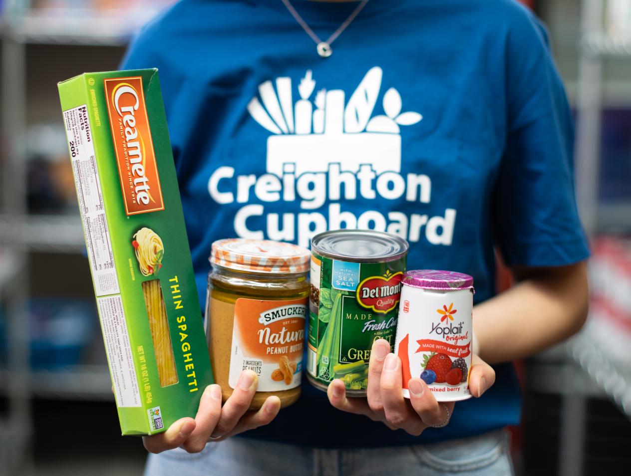 A student wearing a Creighton Cupboard shirt holding donated food items