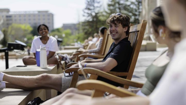 A smiling student with a laptop sitting on a rocking chair outdoors on Creighton's campus.