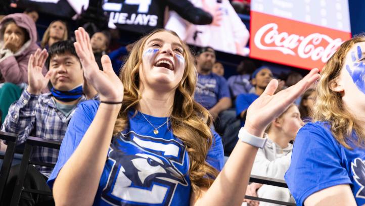 A female student in a blue shirt with face paint cheering at a Creighton sports event, surrounded by a crowd.