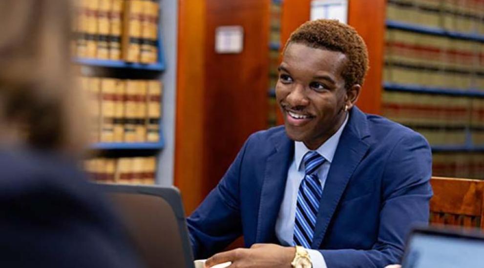 Law student dressed in business suit sitting in law library talking to another student.