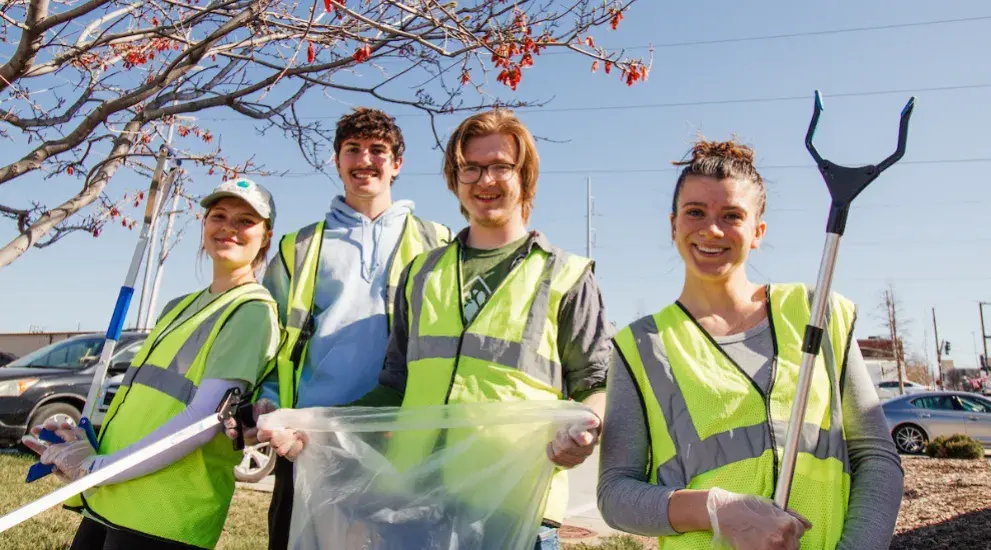 Four students in yellow vest with tools for cleaning up trash.
